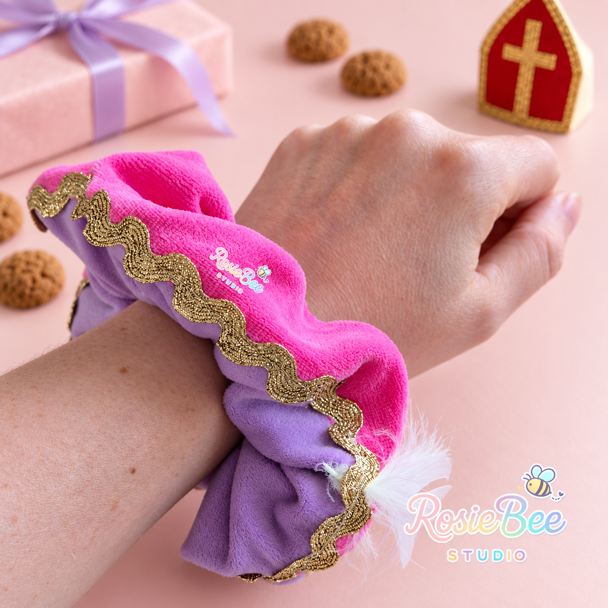 Fuchsia en lila velours pieten scrunchie met gouden golvende sierband en wit veertje, gedragen om een pols. Op de pastelroze achtergrond liggen cadeautjes en kruidnoten naast een rood-gouden mijter.