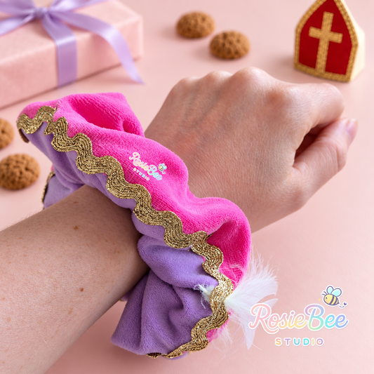 Fuchsia en lila velours pieten scrunchie met gouden golvende sierband en wit veertje, gedragen om een pols. Op de pastelroze achtergrond liggen cadeautjes en kruidnoten naast een rood-gouden mijter.