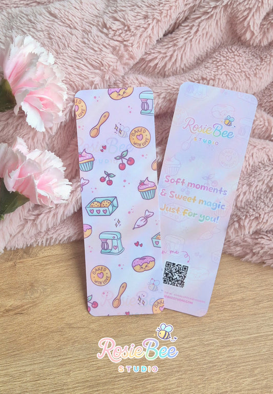 Moji Friends Cosy Café boekenlegger met baked with love bakery patroon en quote achterkant, gefotografeerd op zachte roze deken met bloemen.