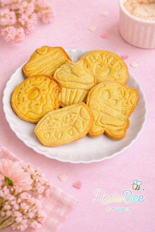 Voorbeeld van gebakken koekjes gemaakt met de Moji Friends Cosy Café cookie cutter set, met cupcake, donut, baked with love, cookie doosje, gebakje en Moji Friend Liefde op een wit bordje in pastelroze setting.