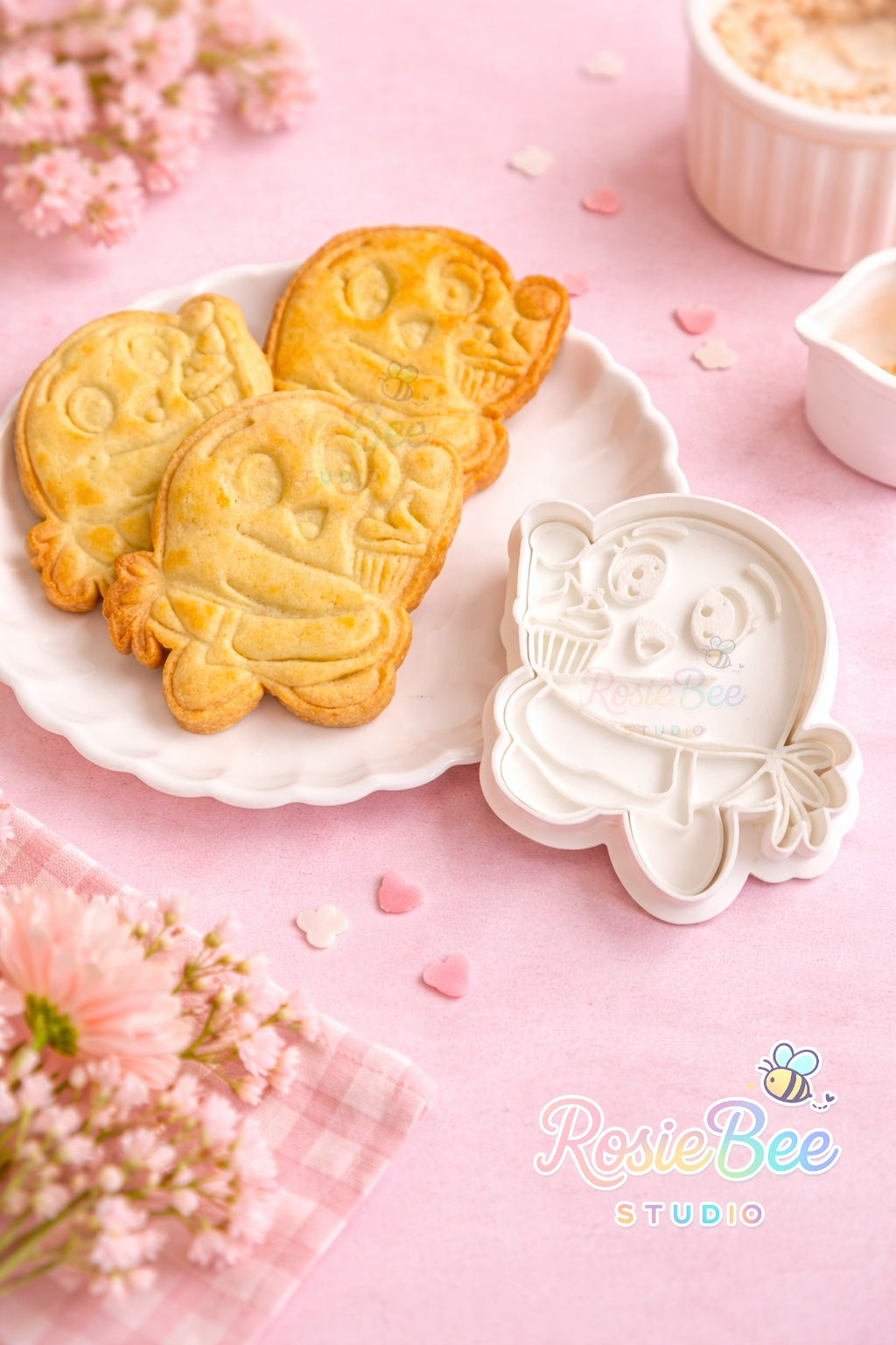 Moji Friend Liefde cupcake koekjesvormpje (cookie cutter) uit de Moji Friends Cosy Café collectie met gebakken koekje op een wit bordje, pastelroze achtergrond en bloemetjes.