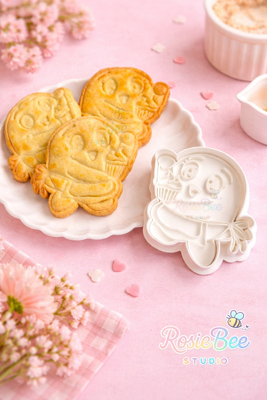 Moji Friend Liefde cupcake koekjesvormpje (cookie cutter) uit de Moji Friends Cosy Café collectie met gebakken koekje op een wit bordje, pastelroze achtergrond en bloemetjes.
