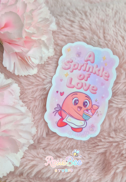 Vinyl sticker “A Sprinkle of Love” met Moji Friend Liefde die een cupcake vasthoudt, gefotografeerd op zachte roze deken met bloemen.