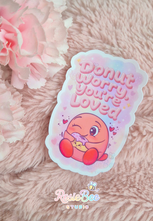Vinyl sticker “Donut Worry, You’re Loved” met Moji Friend Liefde die een donut vasthoudt, gefotografeerd op zachte roze deken met bloemen.