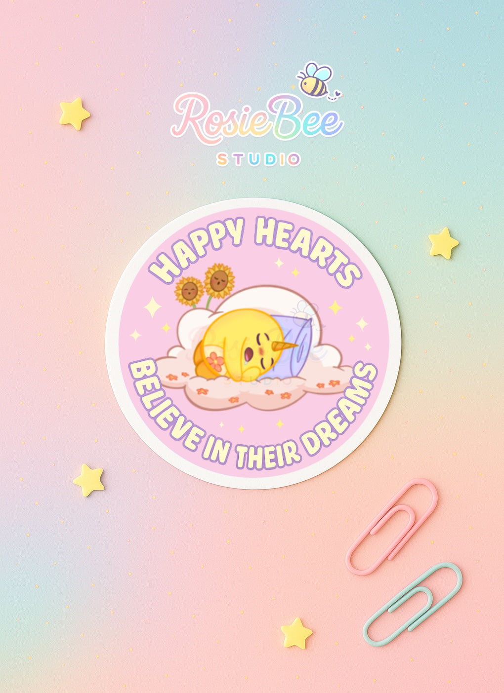 Vinyl sticker “Happy Hearts Believe in Their Dreams” van RosieBee Studio op pastel regenboog achtergrond met sterretjes en paperclips.