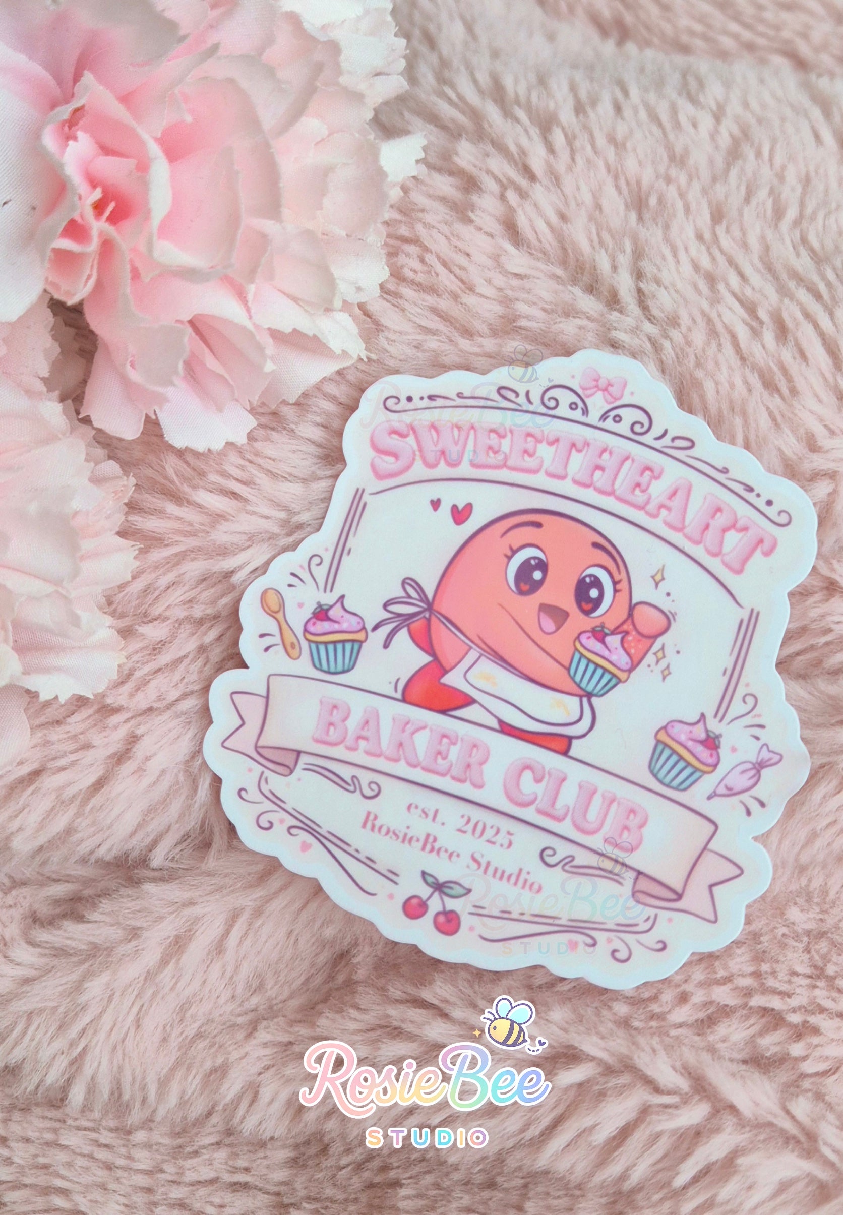 Vinyl sticker “Sweetheart Baker Club” met Moji Friend Liefde in schort met cupcakes, gefotografeerd op zachte roze deken met bloemen.